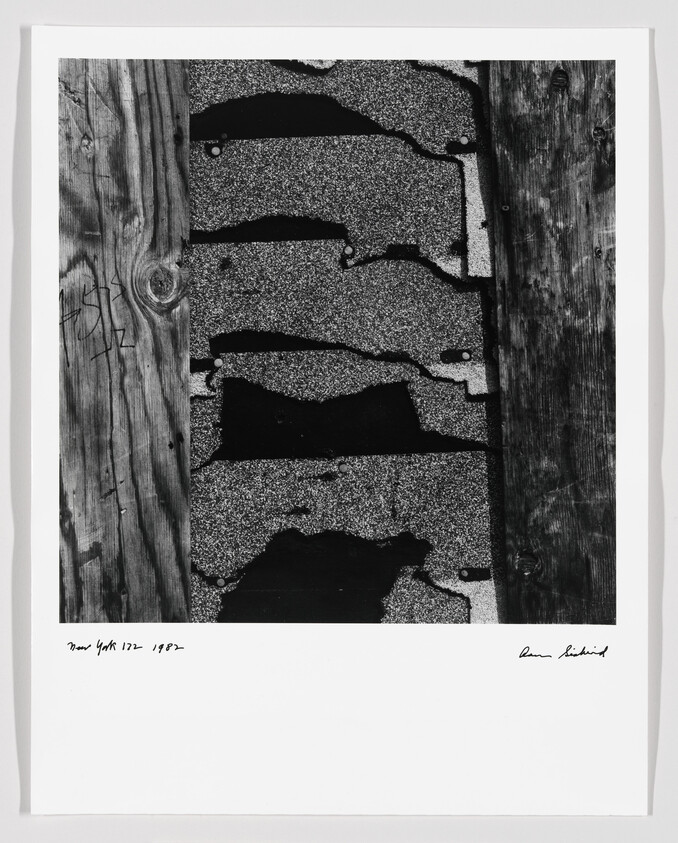 Weathered asphalt shingles with torn edges sandwiched between two rough wooden planks.