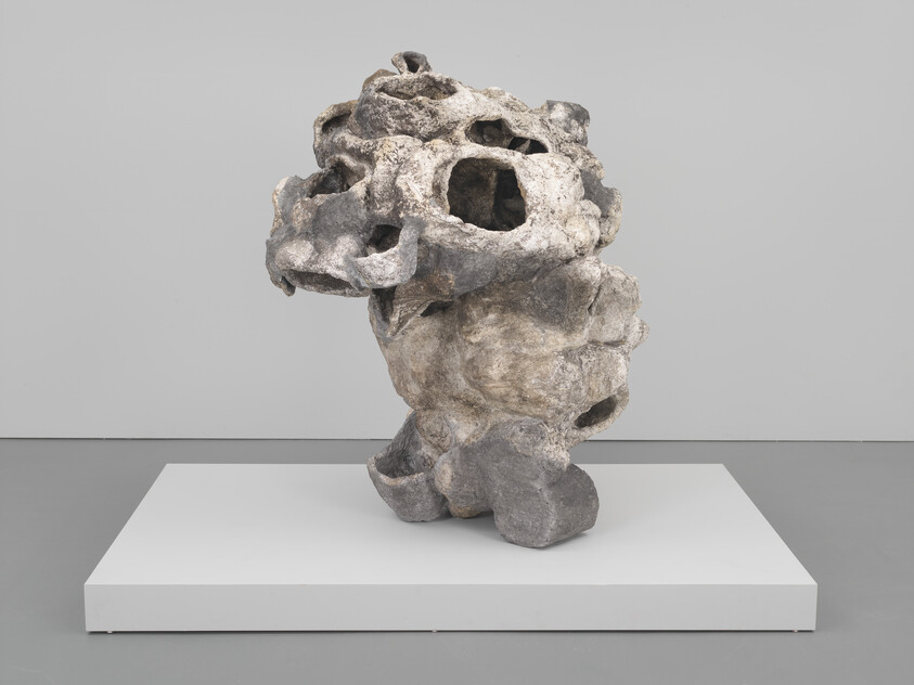 Abstract porous stone sculpture with multiple hollow cavities standing on a white pedestal in a gallery.