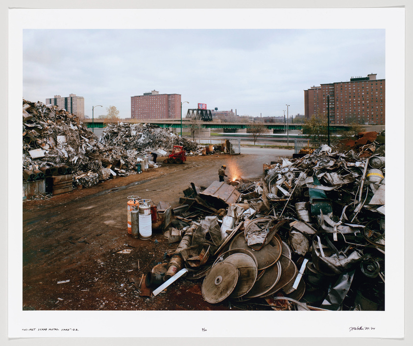 A photograph of a scrap metal yard with large piles of various metal debris, including pieces of machinery, metal scraps, and containers. In the background, there are buildings and a bridge, with overcast skies above. A small fire burns in the middle distance, and the ground appears wet or muddy. The image includes a handwritten caption at the bottom.