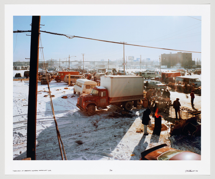 A bustling outdoor market scene with snow on the ground, featuring a variety of vehicles including trucks and cars, and people gathered around stalls and fires for warmth. Power lines and a utility pole frame the left side of the image, and industrial buildings can be seen in the background under a clear blue sky.