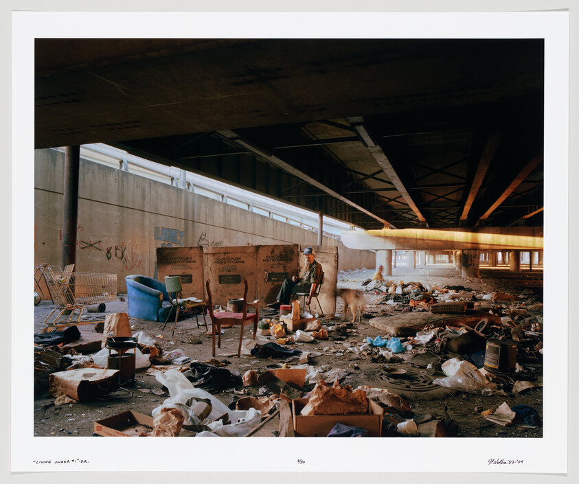 A man and his dog sit among scattered furniture and trash under a highway overpass.