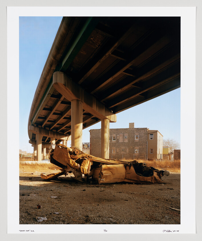 A crushed, rusted car lies under a highway overpass beside empty lots and apartment buildings.