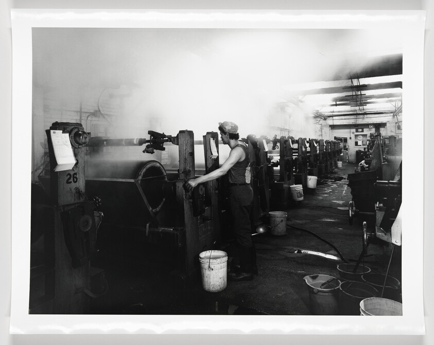 A black and white photo of an industrial setting with a worker operating machinery. The worker is wearing a sleeveless top and a cap, standing next to a large piece of equipment with a wheel and lever. The environment is filled with mist or steam, and multiple similar machines are aligned in rows extending into the background. Buckets, hoses, and industrial debris are scattered across the floor, and paperwork is attached to the machinery in the foreground.