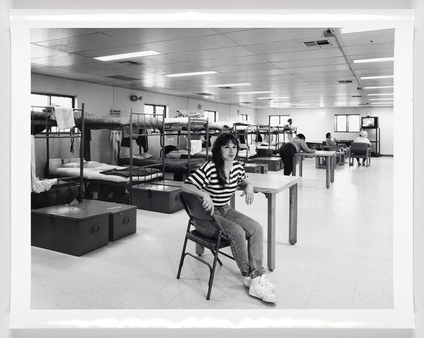 A black and white photo of a young woman sitting on a chair in a communal living space with bunk beds and lockers. She is wearing a striped shirt and jeans, with her hair partially covering her face. In the background, other individuals are seated at tables, and one person is watching a television.