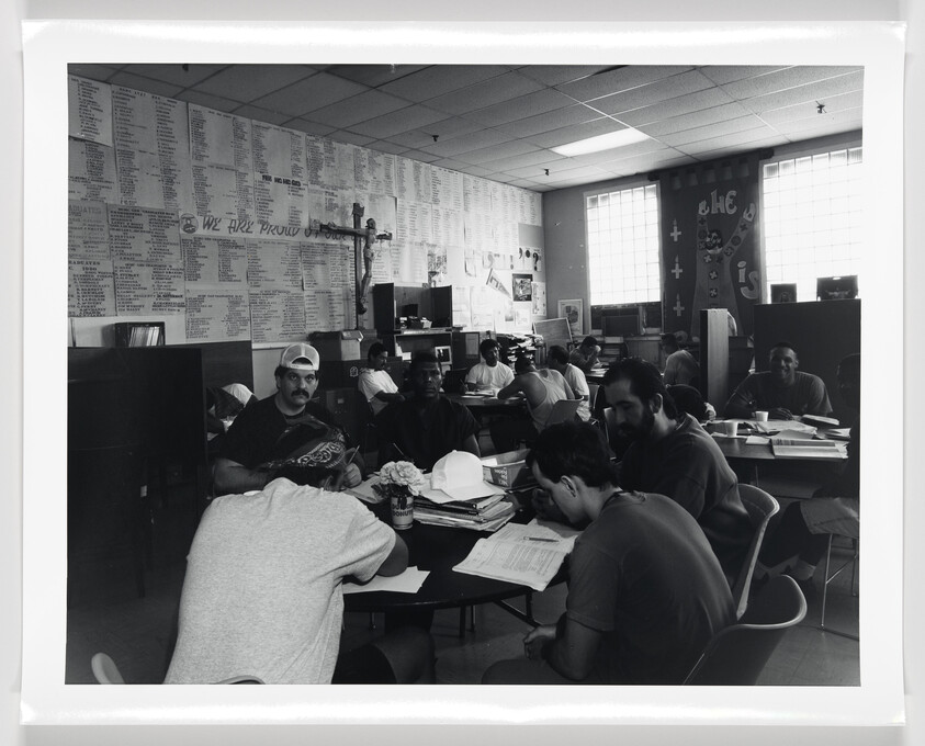 Several people sit at tables in a classroom, reading and writing on papers, with names listed on the wall.