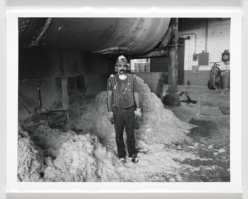 A black and white photograph of a man standing in an industrial setting with a large pile of a cotton-like substance around him. He is wearing a patterned shirt, pants covered in the substance, and a protective mask on his head. There are shovels and a wheelbarrow nearby, suggesting manual labor. The environment looks dusty and there's another person visible in the background.