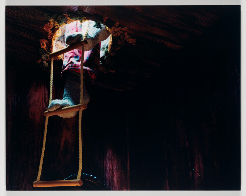 A person in socks climbs a rope ladder up through a rough hole in a wooden ceiling.