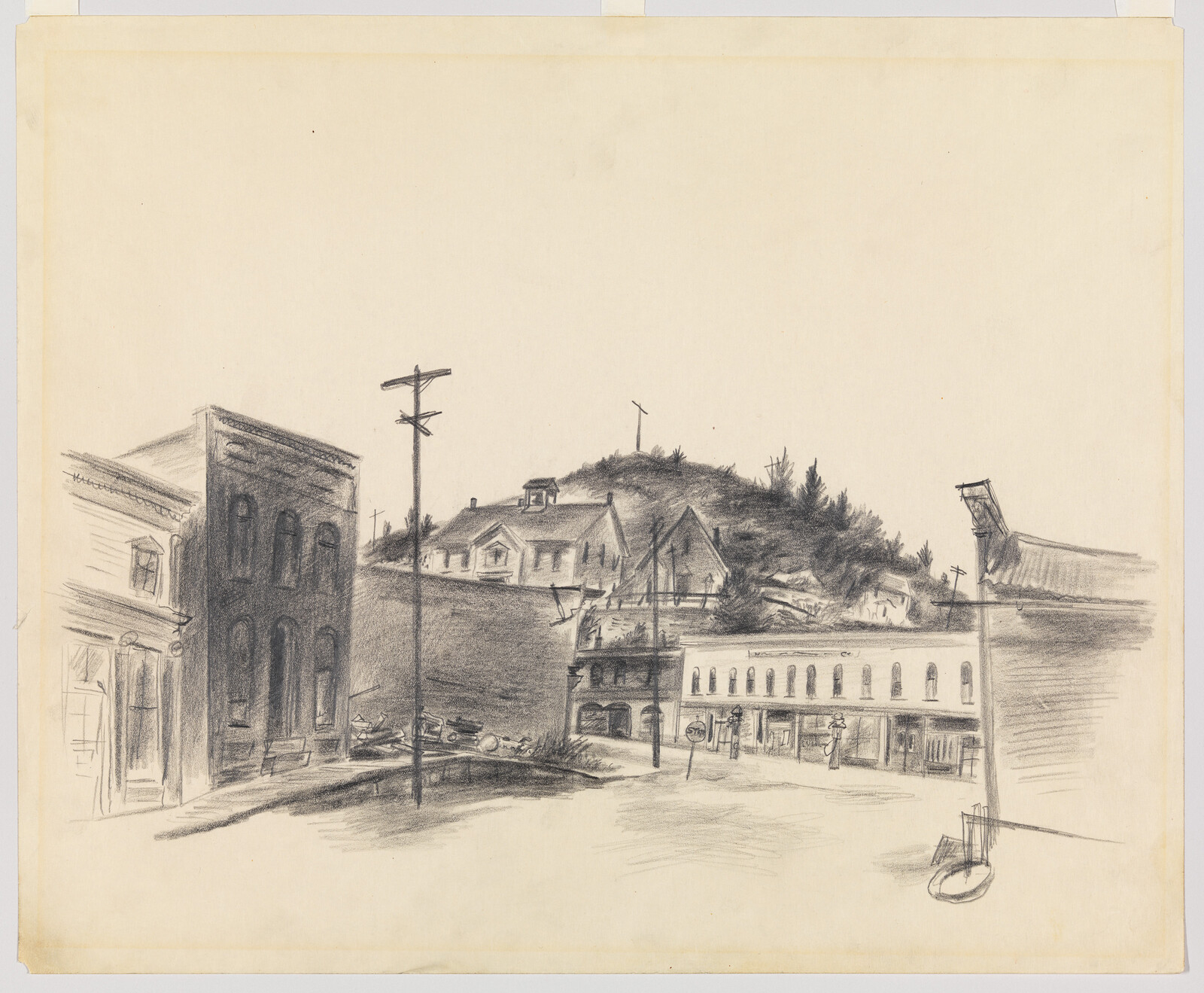 Small town street with storefronts, telephone poles, and houses on a wooded hill in the background.