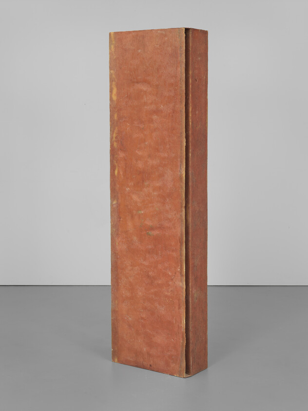 A tall, narrow, rectangular sculpture with a rust-colored surface stands upright against a white wall on a grey floor.