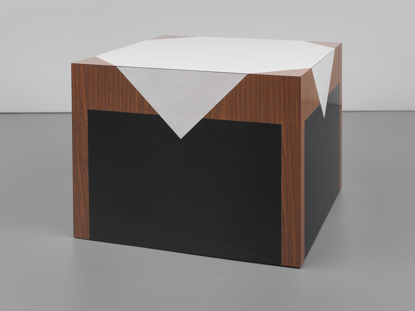 A cube table with wood veneer edges, black panels, and a white cloth draped over the top.