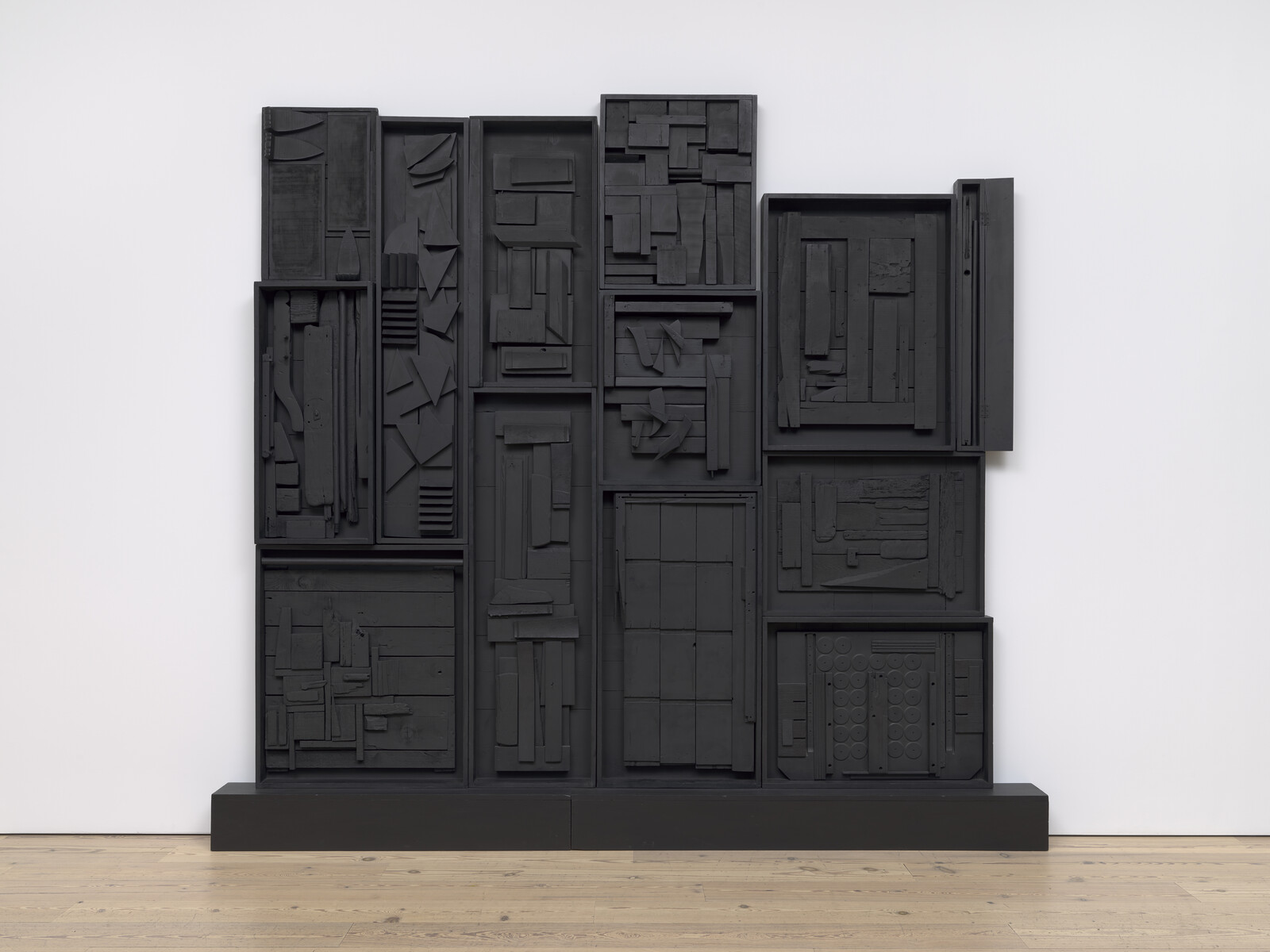 Black abstract wall sculpture with various geometric shapes and textures, displayed on a wooden floor.