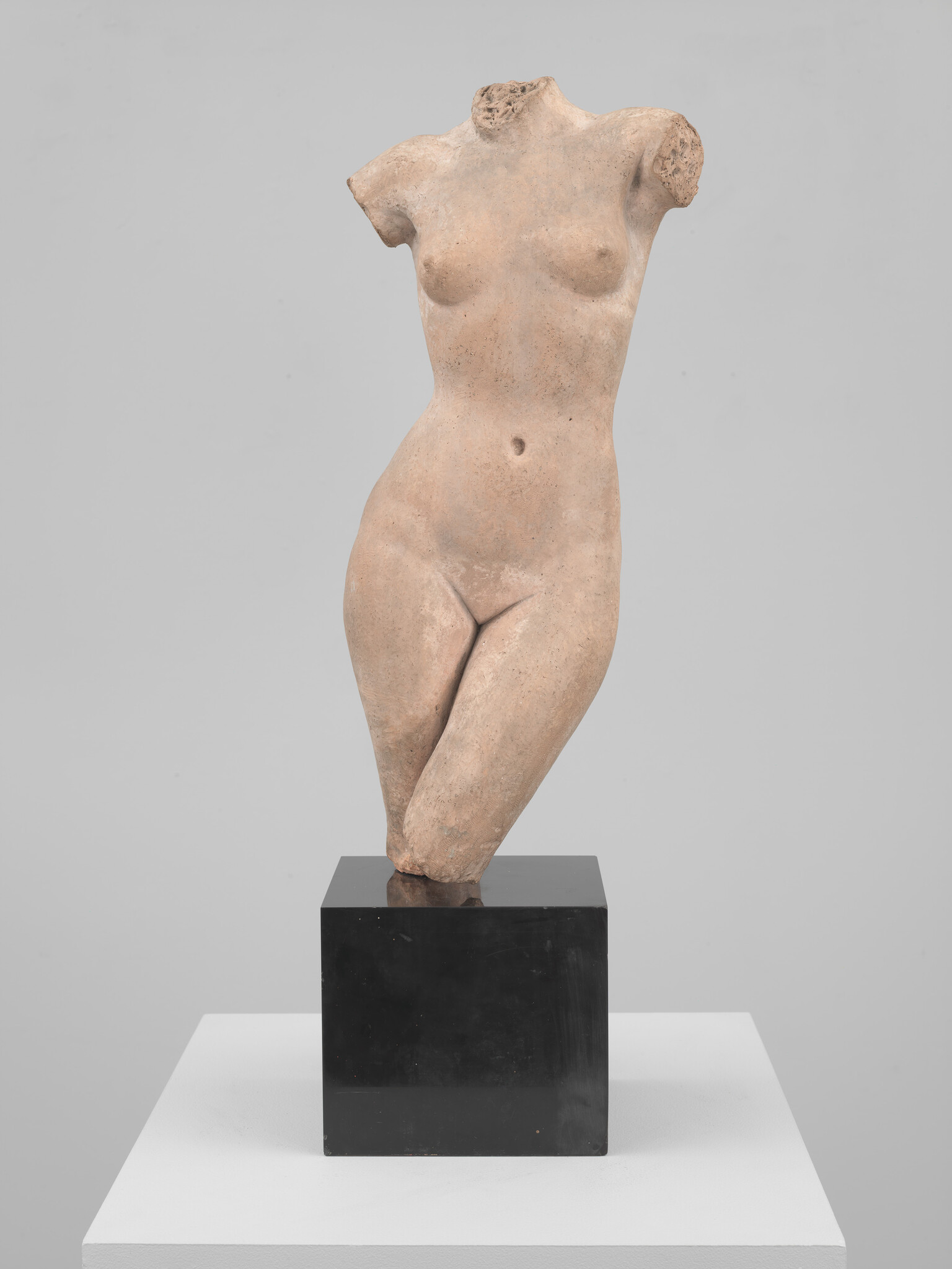A fragmented classical female torso sculpture stands on a black pedestal in a neutral gallery.