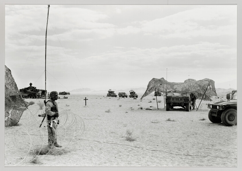 A soldier stands by concertina wire in a desert camp near military vehicles and camo netting.