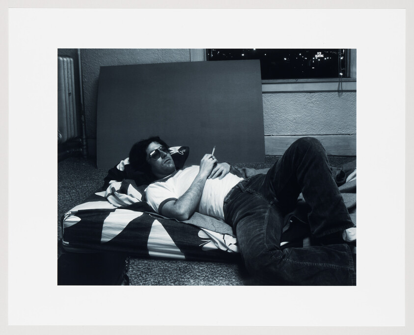 A black and white photo of a man lying on his side on a striped mattress on the floor, wearing sunglasses, a white t-shirt, and jeans. He is holding a cigarette in one hand and appears relaxed. There's a large blank canvas leaning against the wall behind him, and a window showing a night scene with a few lights in the distance.