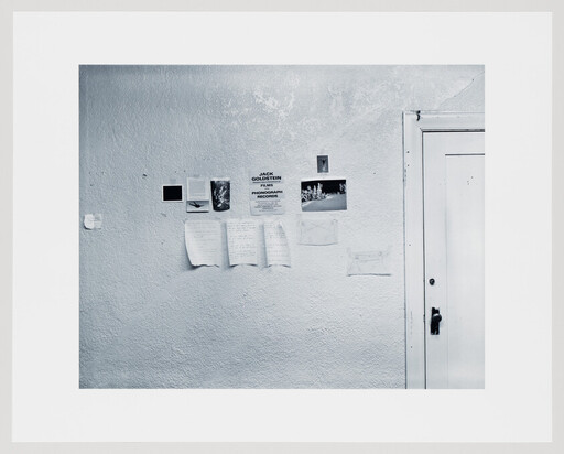 A textured white wall displays several taped photos and handwritten notes beside a closed white door.