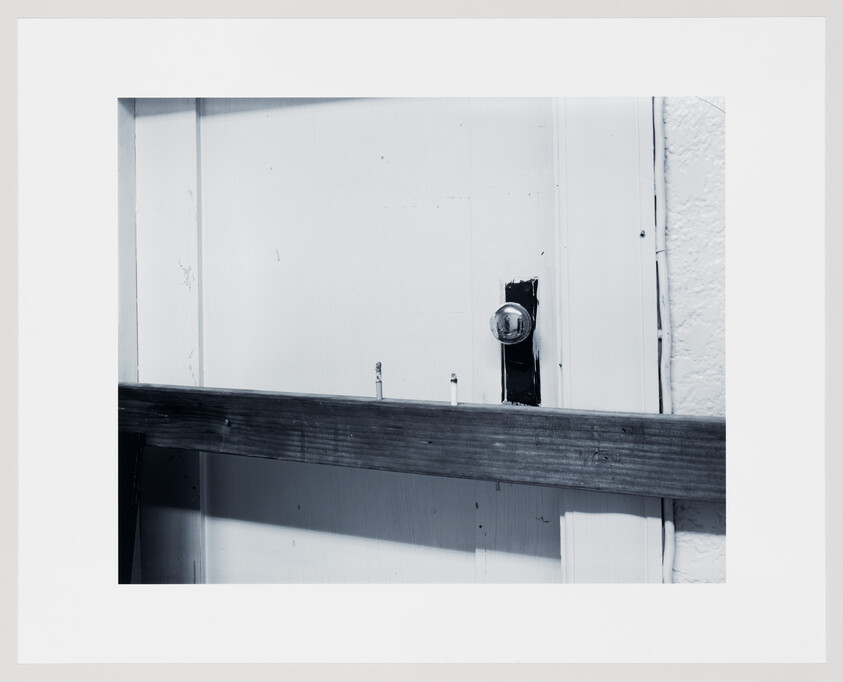 A white door is barricaded with a horizontal wooden plank nailed across near a round glass doorknob.