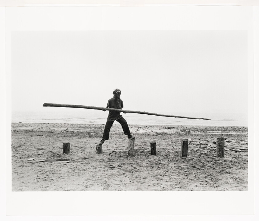 A person balances across wooden stumps on a beach while holding a long pole.