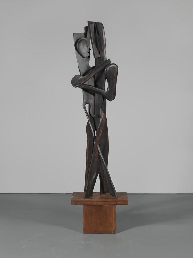Tall abstract wooden sculpture of two elongated figures embracing on a square wooden pedestal.