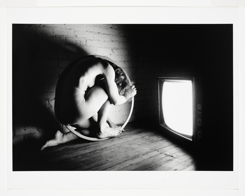 Naked person curled inside a circular hoop on the floor facing a glowing television.