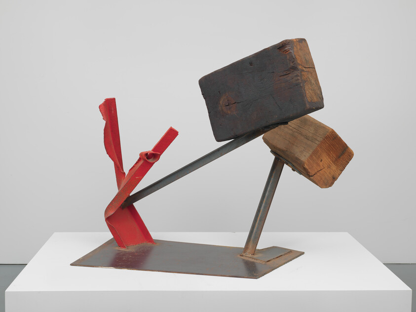 Abstract metal and wood sculpture with two large wooden blocks balanced on angled steel supports above a red base.