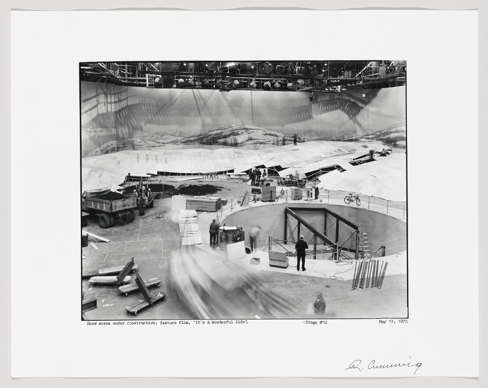 Crew members build a large indoor snow scene set with backdrop and equipment on a film stage.