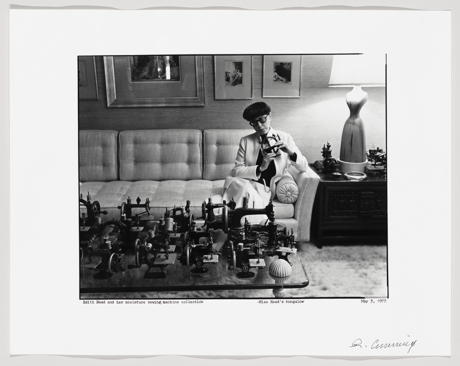 A black and white photograph depicting a woman sitting on a couch, examining a miniature sewing machine from a collection displayed on a coffee table in front of her. The setting appears to be a cozy room with a framed picture on the wall, a lamp, and other decor items. The photo includes captions indicating the subject as Edith Head and the location as her bungalow, dated May 5, 1977, with a signature at the bottom right.