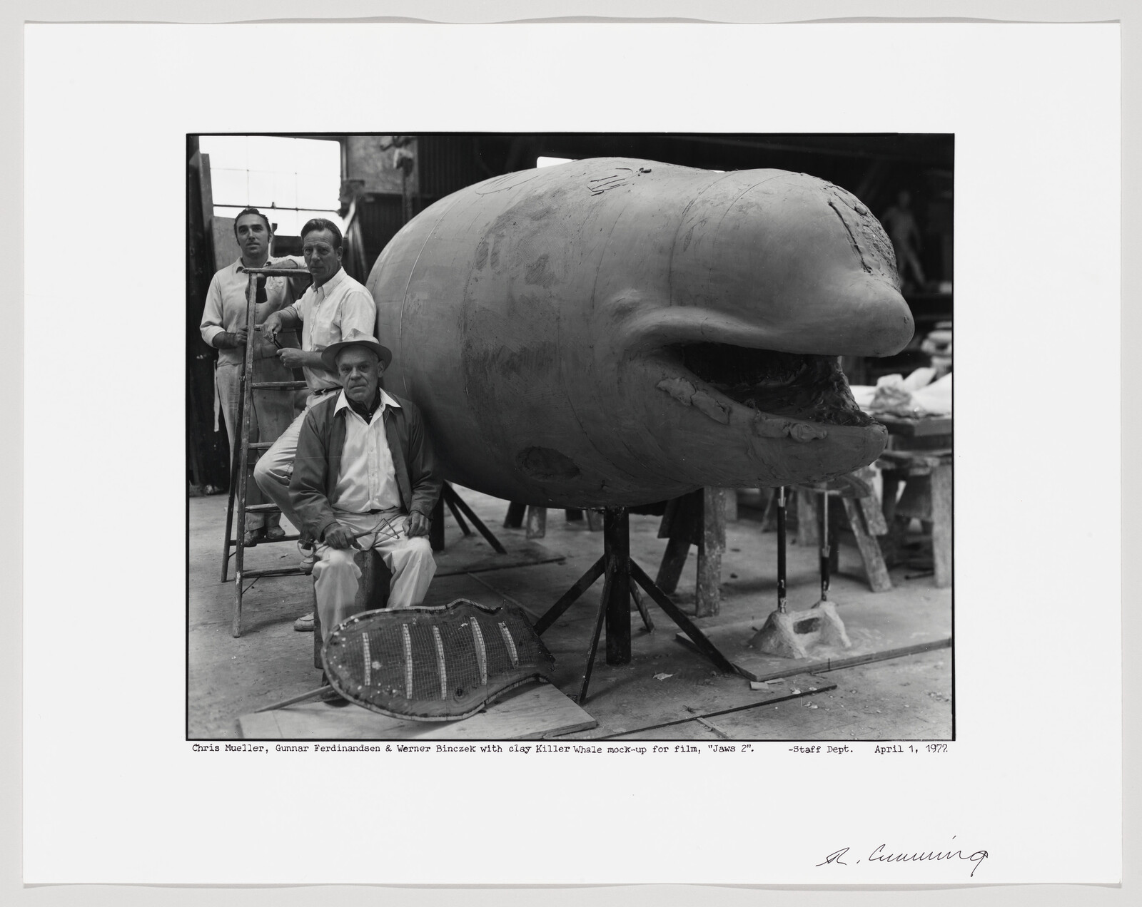 A black and white photo showing three men posing with a large clay model of a killer whale, which is a mock-up for the film "Jaws 2". One man is seated in front of the model, while the other two stand behind it. The model is supported by a metal frame and the photo includes a caption with the names of the men and the date, April 11, 1977, along with a signature in the bottom right corner.