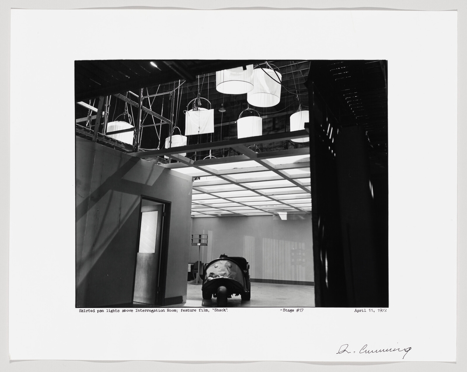 Small prop car parked under lit ceiling grid on a film set with hanging lights.