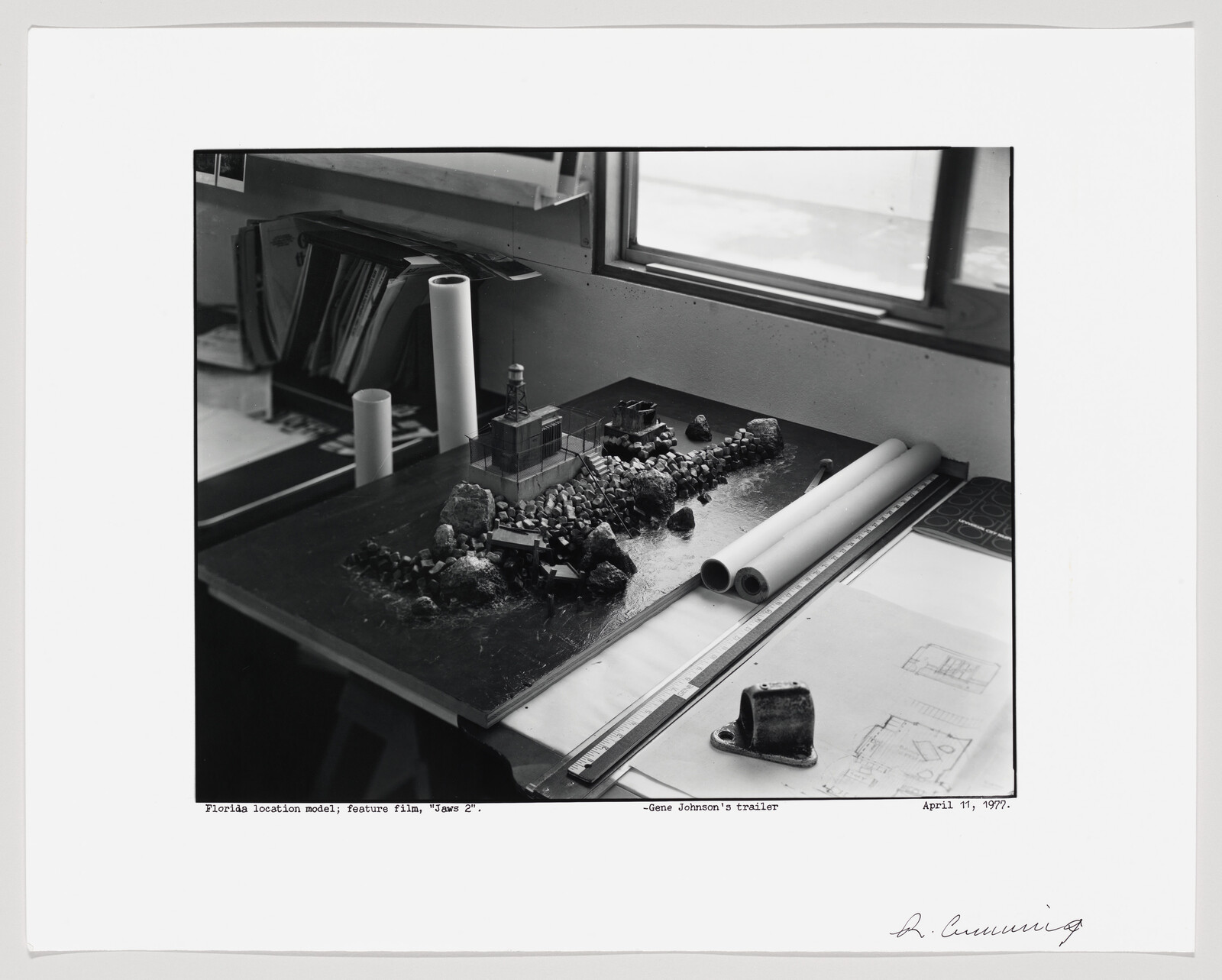 A black and white photograph of a work desk with a miniature building model and surrounding landscape, rolls of paper, and architectural drawings. The image includes handwritten notes and a date at the bottom, suggesting an artistic or architectural project.