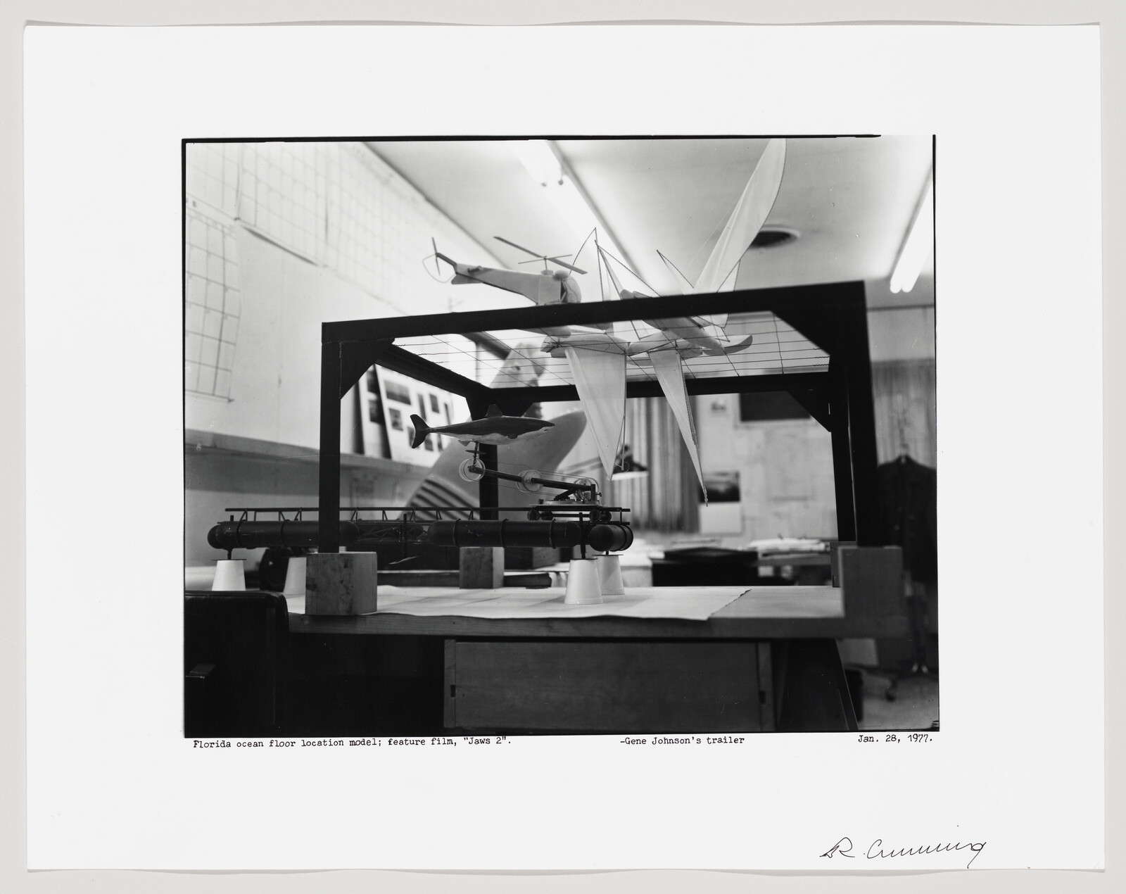 A black and white photograph of a model setup for the ocean floor, presumably used in the feature film "Jaws 2." The model includes a miniature boat on a simulated water surface, with various other elements that suggest an underwater scene. The photo is taken in a studio environment with tables, papers, and other materials visible in the background. The image is dated January 28, 1977, and includes a caption at the bottom.