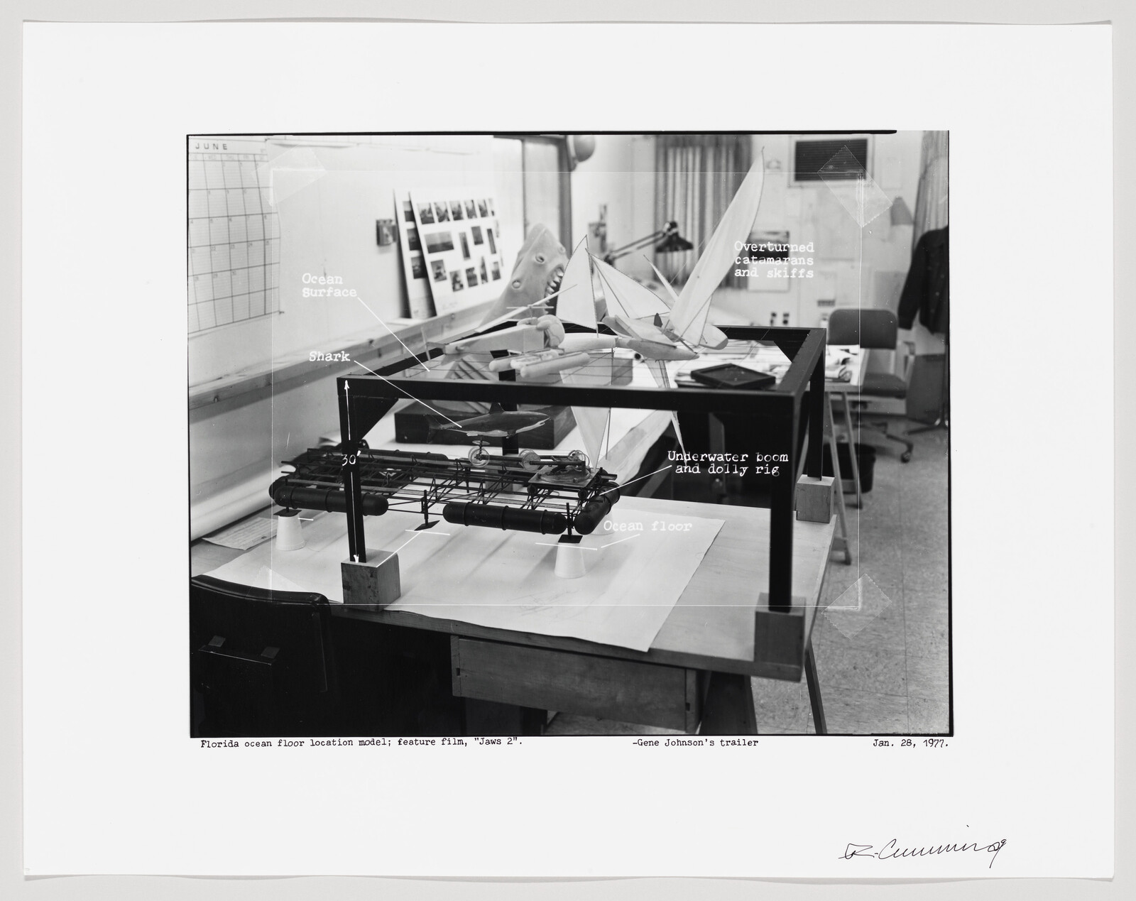 Black and white photo of a scale model setup for the film "Jaws 2," showing a miniature ocean surface and ocean floor with labels such as "Shark," "Overturned catamarans and skiffs," and "Underwater boom and dolly rig." The model is in a studio environment with various equipment and a calendar on the wall, dated January 28, 1977.
