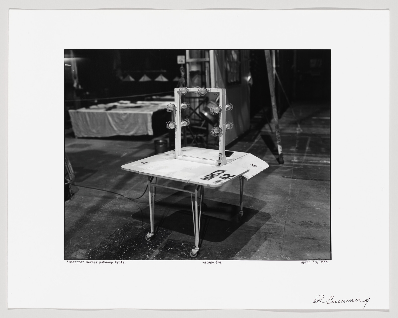 A black and white photograph depicting a makeup table with a mirror surrounded by light bulbs, presumably from the "Barretti" series, situated on a stage. The table is cluttered with various items and there's a sign that reads "WET PAINT" on it. The background shows a partially visible draped table and a ladder, indicating a behind-the-scenes setting. The image includes handwritten text and a signature at the bottom, along with a date: April 18, 1977.
