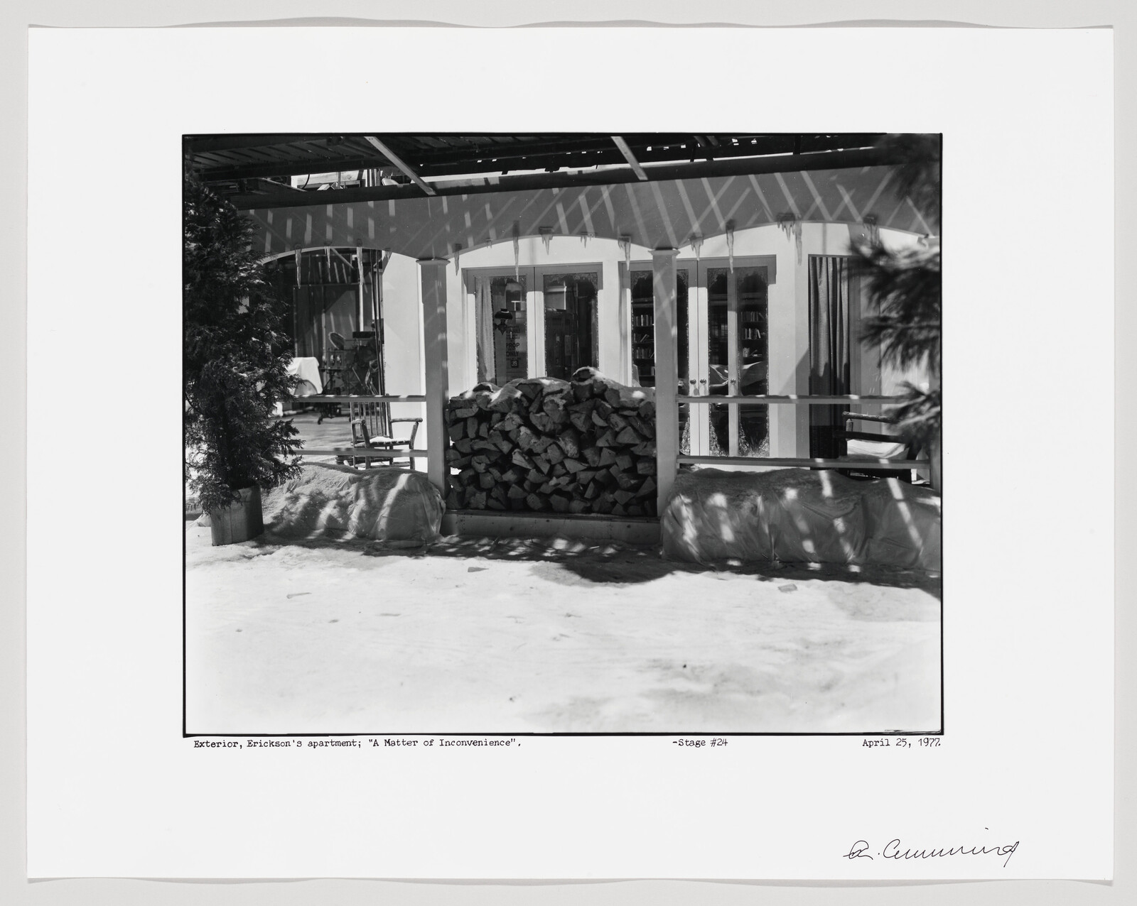 Black and white photograph of the exterior of Erickson's apartment with a caption "A Matter of Inconvenience" and a date "April 25, 1972". The image shows a neatly stacked pile of firewood next to a porch with a snow-covered ground, a bench, and a part of a tree visible on the left. Shadows from the porch railing create patterns on the snowy surface.