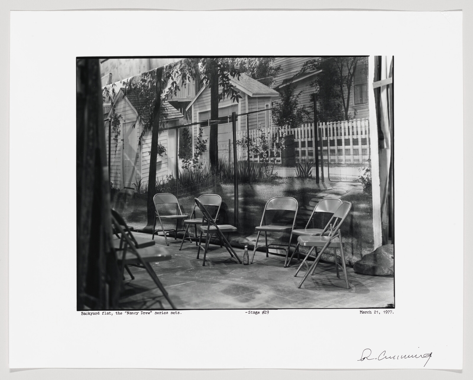 Black and white photograph of a staged outdoor scene with four metal folding chairs in the foreground, set on a concrete floor. The background features a painted backdrop depicting a residential setting with a white picket fence, a house, and lush vegetation. The scene is labeled as a set for the "Nancy Drew" series, located on stage #29, and is dated March 21, 1977, with a signature at the bottom right.