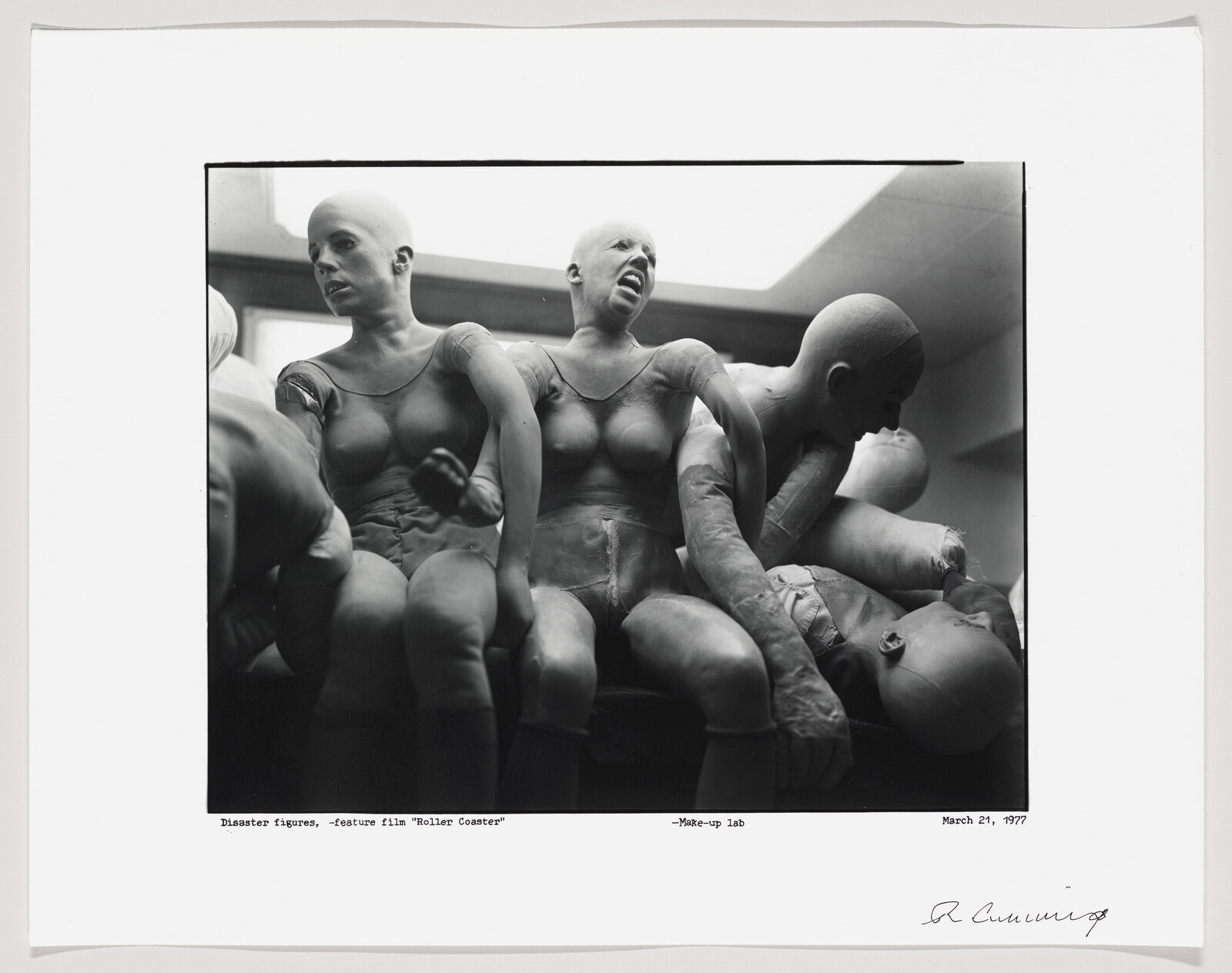 A black and white photograph showing a collection of mannequin-like figures with exaggerated expressions and poses, suggesting distress or disaster. The figures are bald and appear to be in a make-up lab, as indicated by the caption, which also references the feature film "Roller Coaster." The photo is dated March 21, 1977, and signed by R. Cummings.