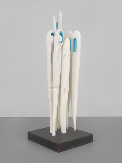 Tall white wooden sculpture with elongated figures and blue interior niches on a black base.