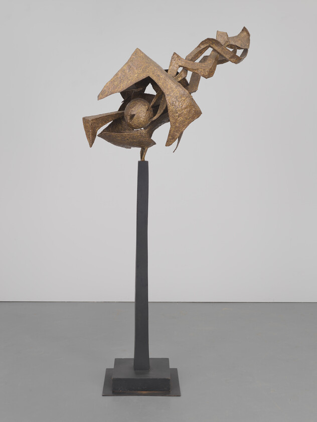 A modern abstract metal sculpture mounted on a tall, slender pedestal. The sculpture features geometric shapes and forms that appear to be dynamically arranged, with a rusted texture, giving it an industrial look. The background is a plain, light-colored wall on a grey floor.