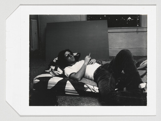 A man wearing sunglasses lies on a striped mattress on the floor smoking a cigarette.