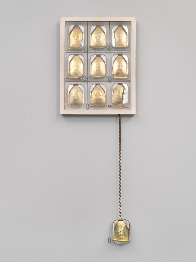 A framed nine-tile grid of gold metal plates with one plate hanging by a chain.