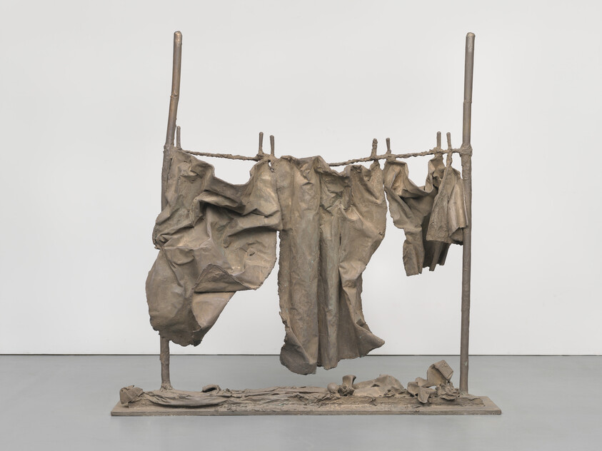Sculpture of weathered clothes hanging on a rope between two poles, with crumpled fabric below.