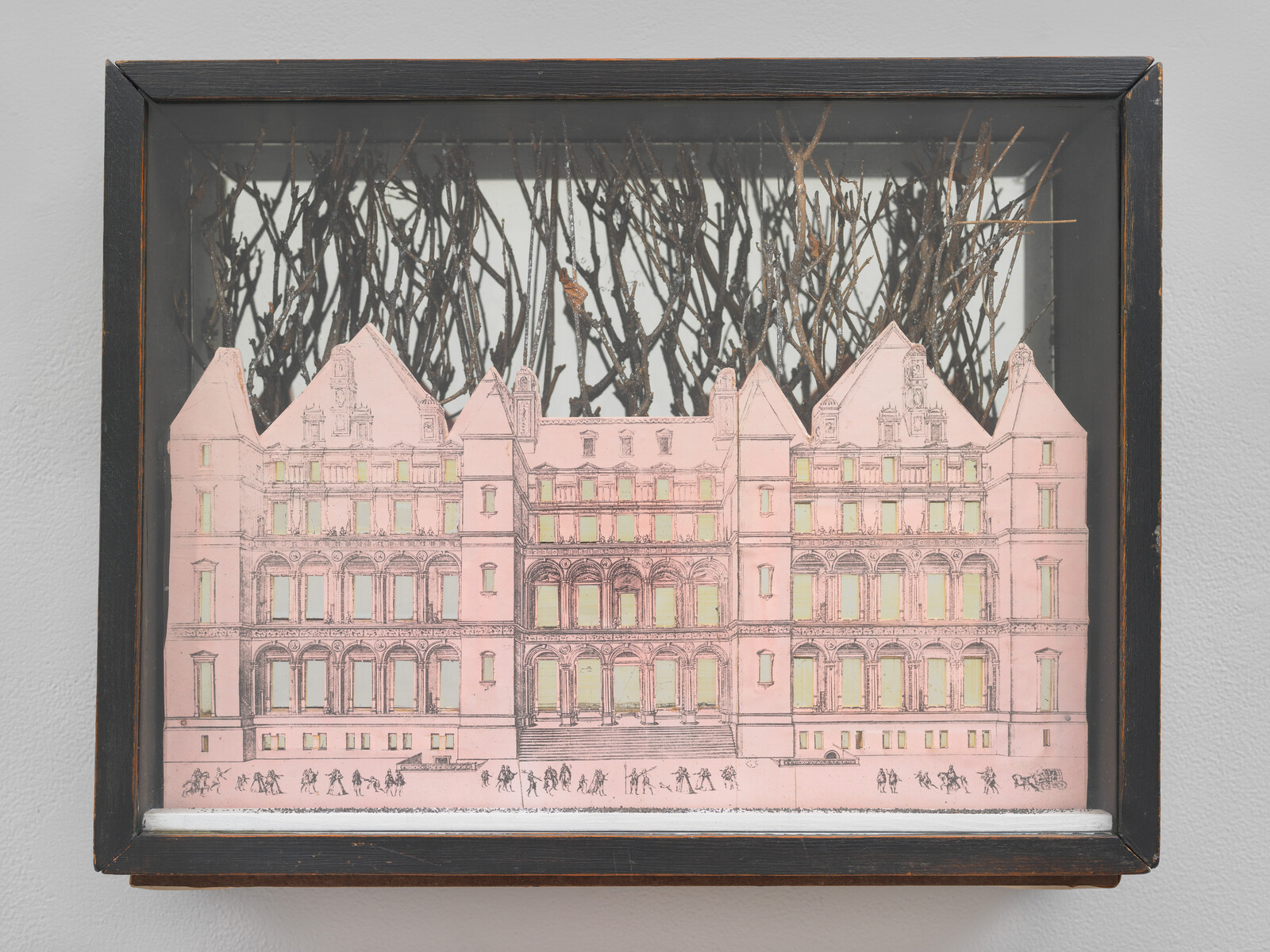 Framed shadowbox contains a pink paper cutout of a large building with twigs behind it.