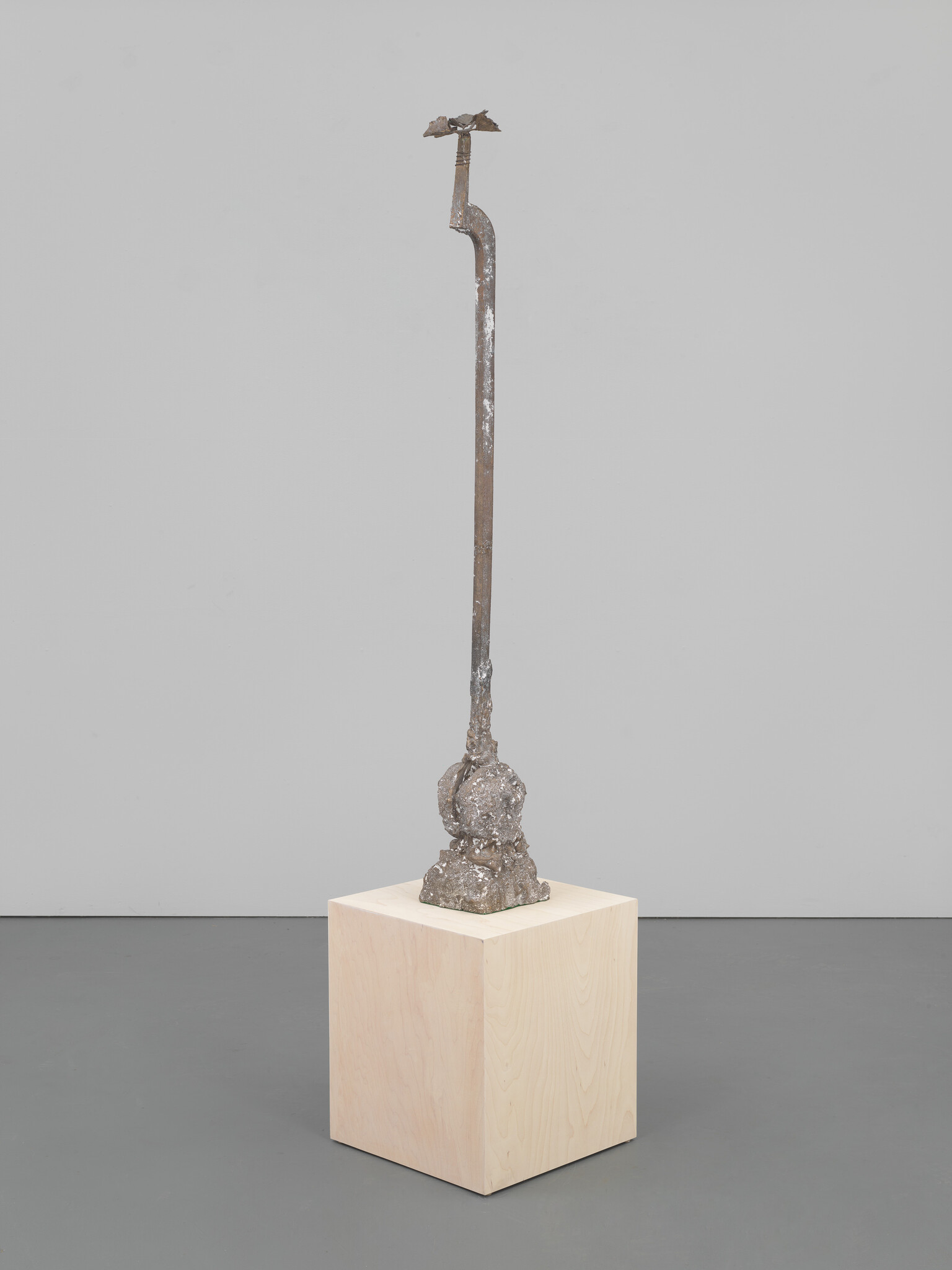A tall rusted metal rod sculpture with a rough, lumpy base displayed on a wooden plinth.