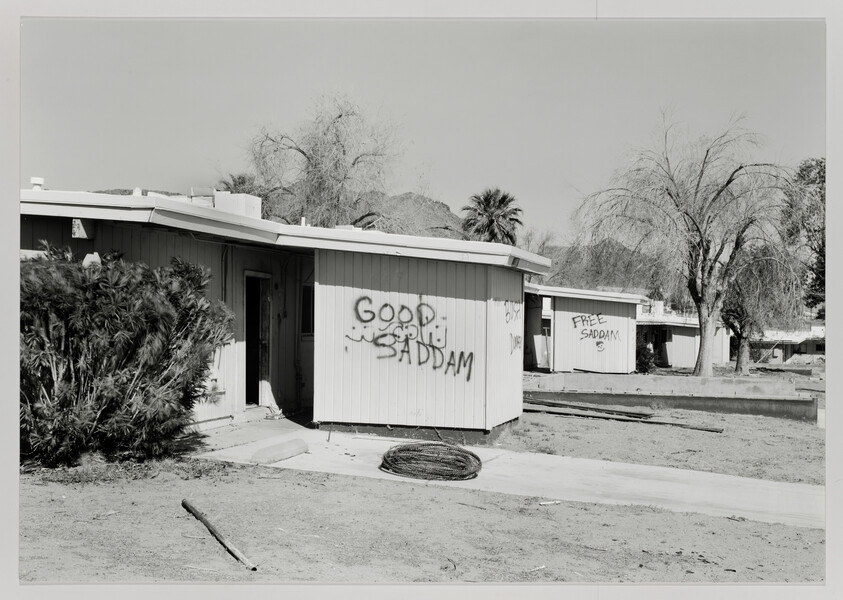Single-story homes in a desert yard have graffiti reading "GOOD SADDAM" and "FREE SADDAM" on their walls.