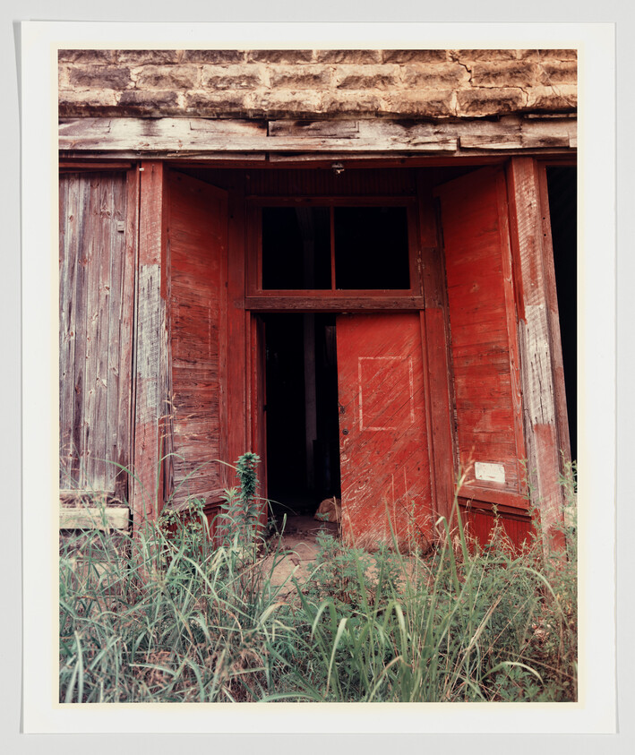 A weathered red wooden door set in a rustic building with a stone lintel above it. The door is partially open, revealing a dark interior, and tall grasses grow in the foreground.