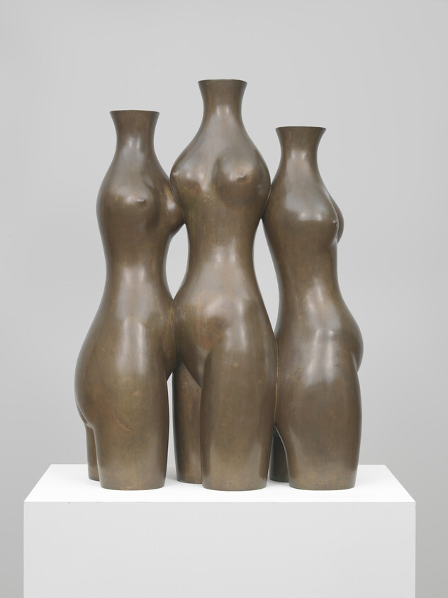 Three headless bronze female torsos standing closely together on a white pedestal.