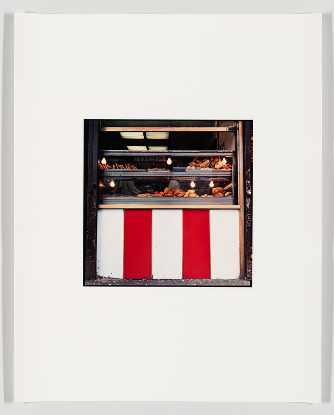 A framed photograph of a bakery window displaying an assortment of bread and pastries, with a red and white striped panel below the window.