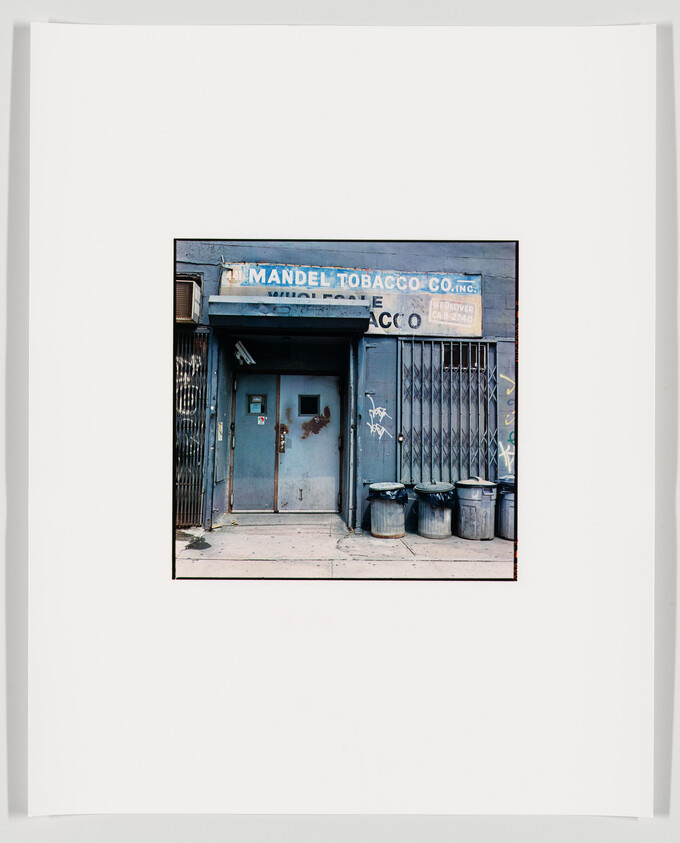 A photograph of an old, weathered storefront with a sign reading "Mandel Tobacco Co. Inc." above a closed, graffiti-tagged metal gate and a door with peeling blue paint. Three trash cans are lined up against the wall to the right of the door. The image is mounted on a white background with a wide border.