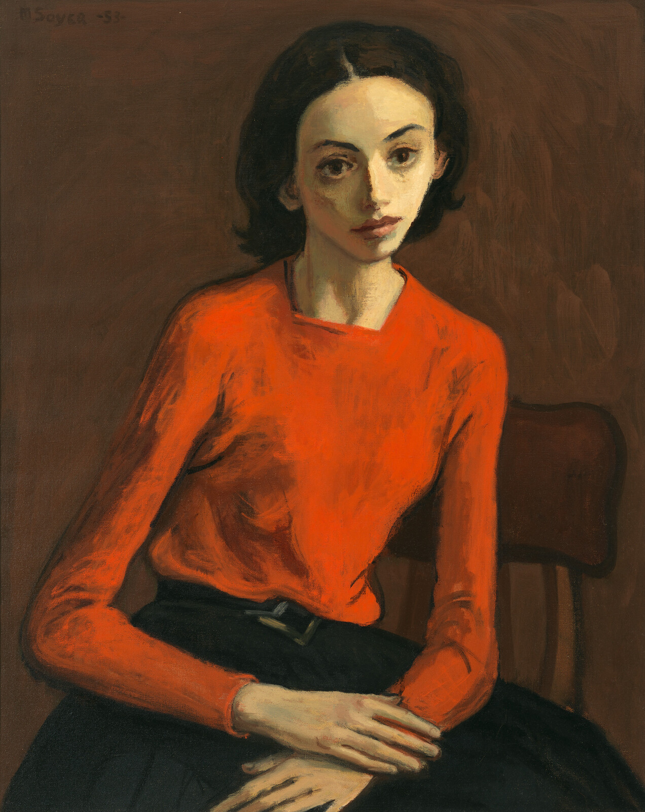 Young woman in an orange sweater sits on a chair with hands folded in her lap.