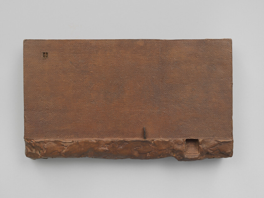 Rectangular brown textured sculpture with a rough base and a small recessed opening on the right.