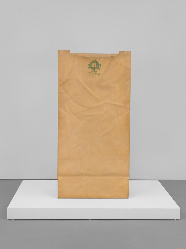 Tall brown paper bag standing upright on a white platform against a gray wall.